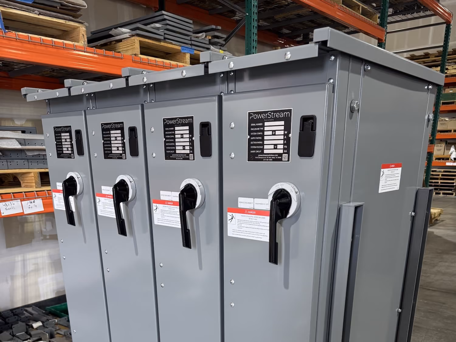 Four gray PowerStream electrical combiner boxes with black handles mounted side by side in a warehouse with shelves and industrial equipment.