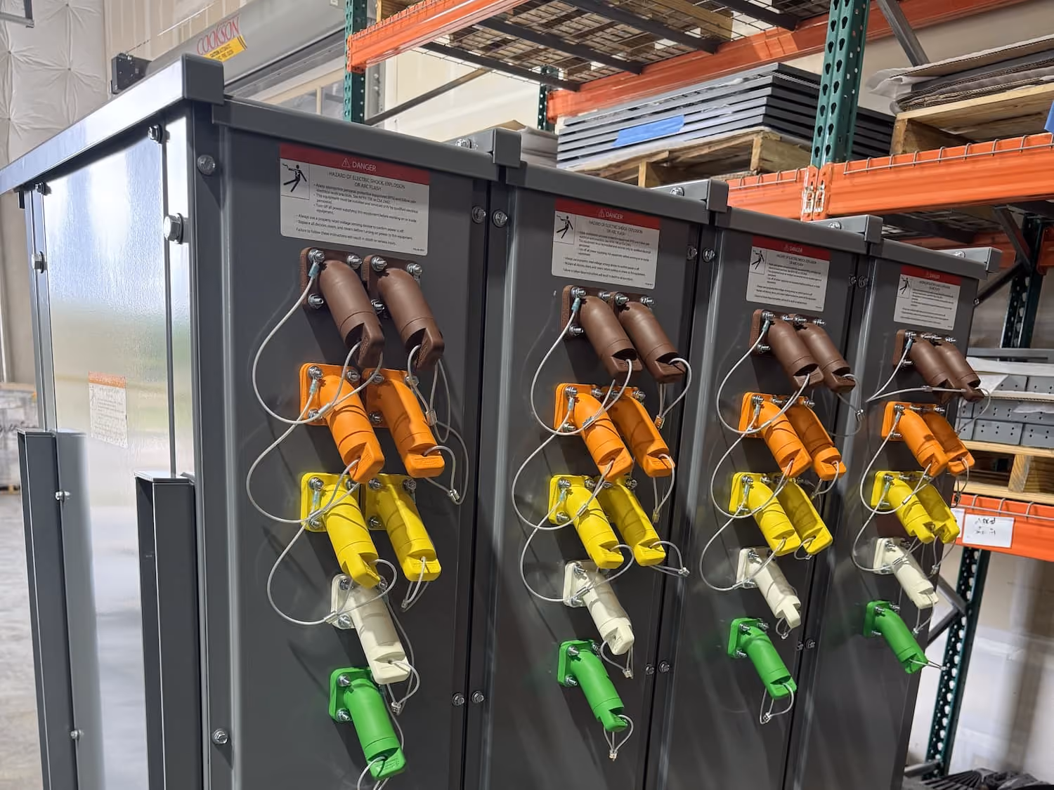 Four identical gray electrical combiner boxes with color-coded insulated handles in brown, orange, yellow, white, and green, mounted in a row inside a warehouse with metal shelving.