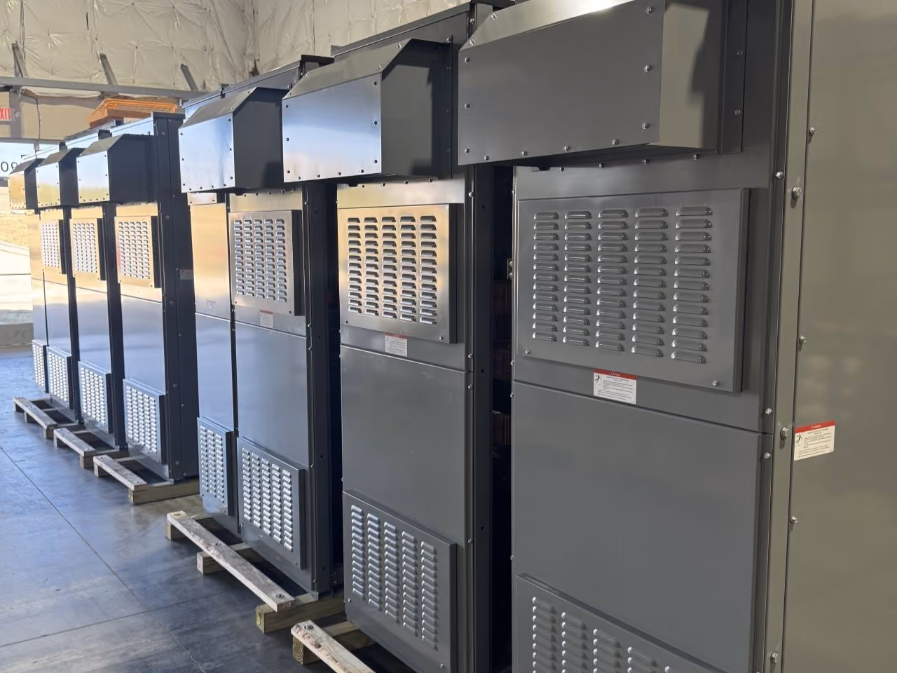 Three large industrial metal enclosures with vented panels and protective covers, placed side by side on wooden pallets inside a warehouse with insulation on the walls.