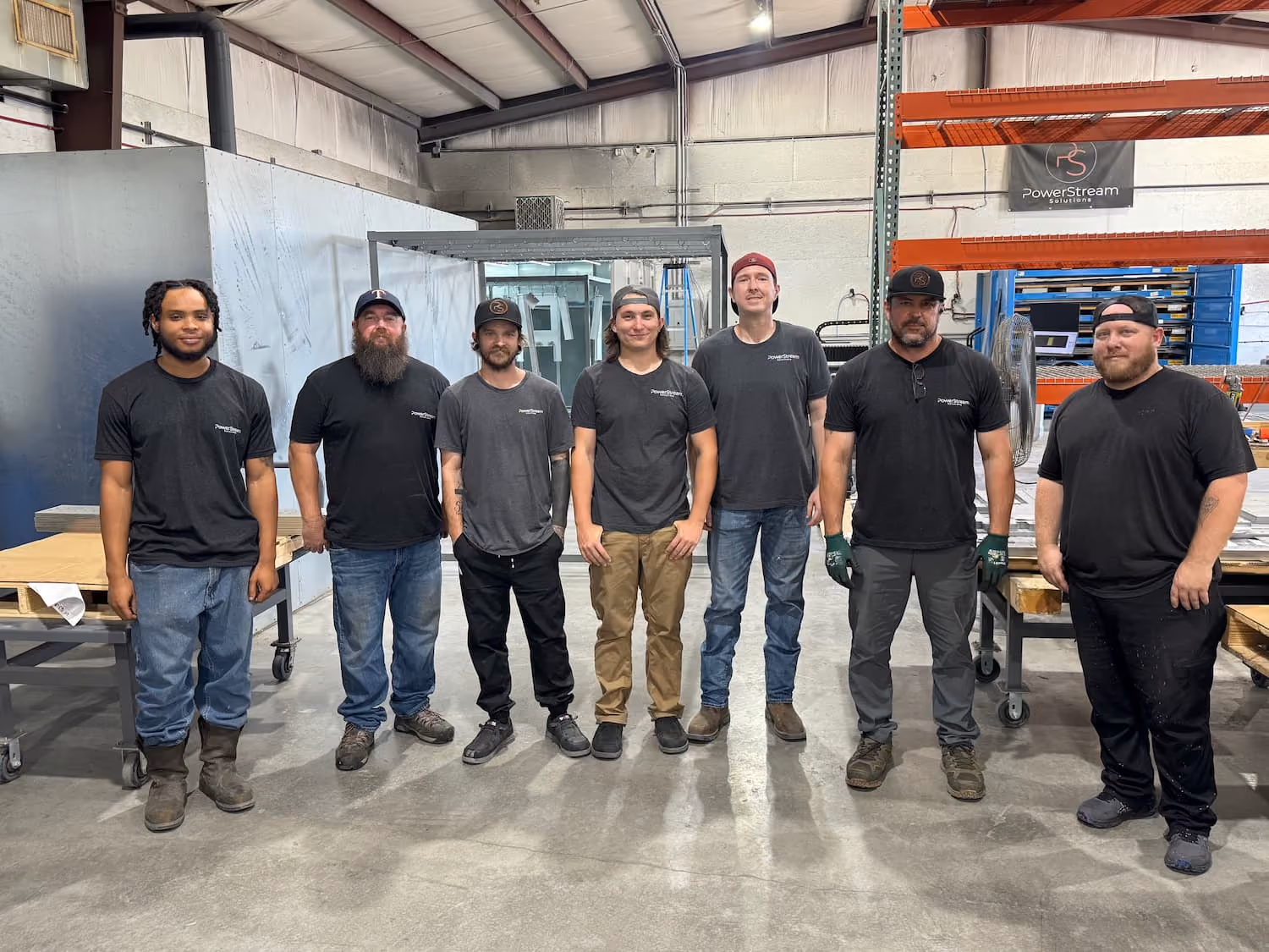 Seven men standing in a warehouse or workshop, all wearing dark shirts and casual work pants, with industrial equipment and shelves visible in the background.