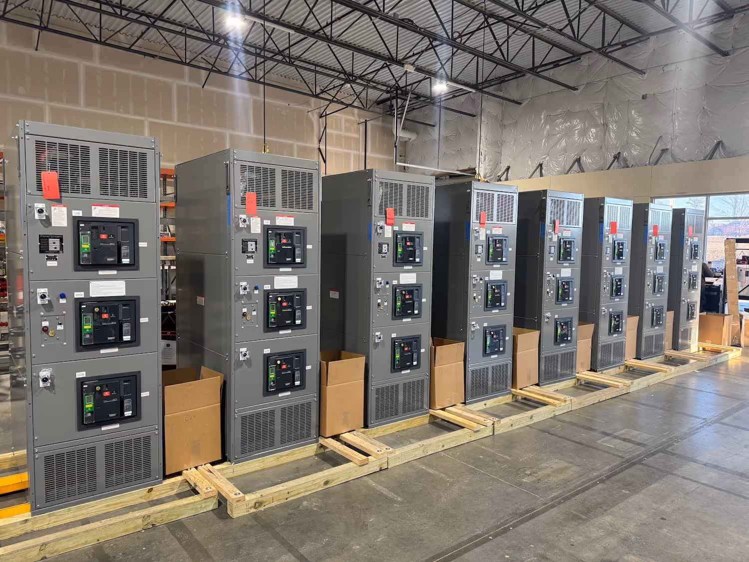 Row of large industrial electrical panels or circuit breaker cabinets lined up inside a warehouse with a high ceiling and concrete floor.