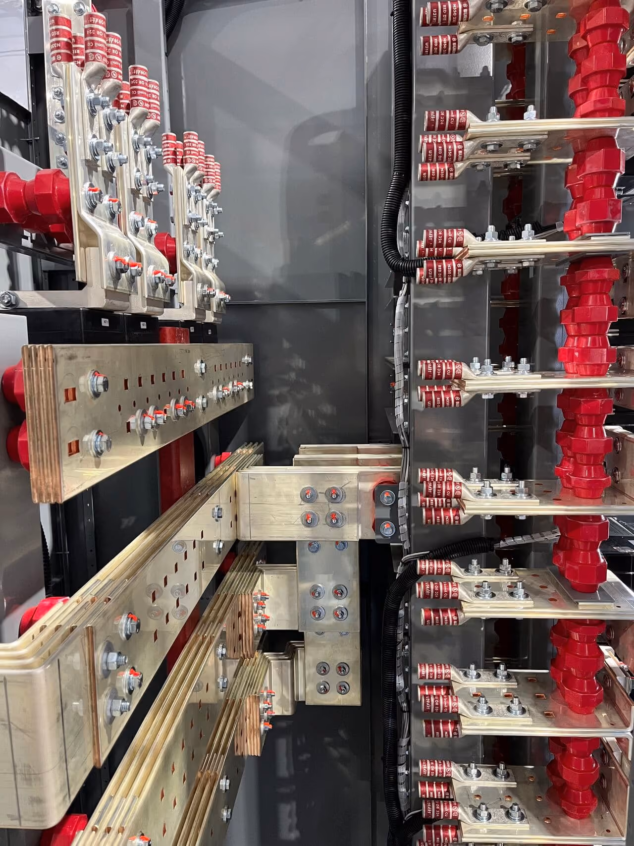 Close-up of power distribution busbars inside an electrical panel, showing metal connectors, numerous bolts, and red insulating components arranged vertically and horizontally.