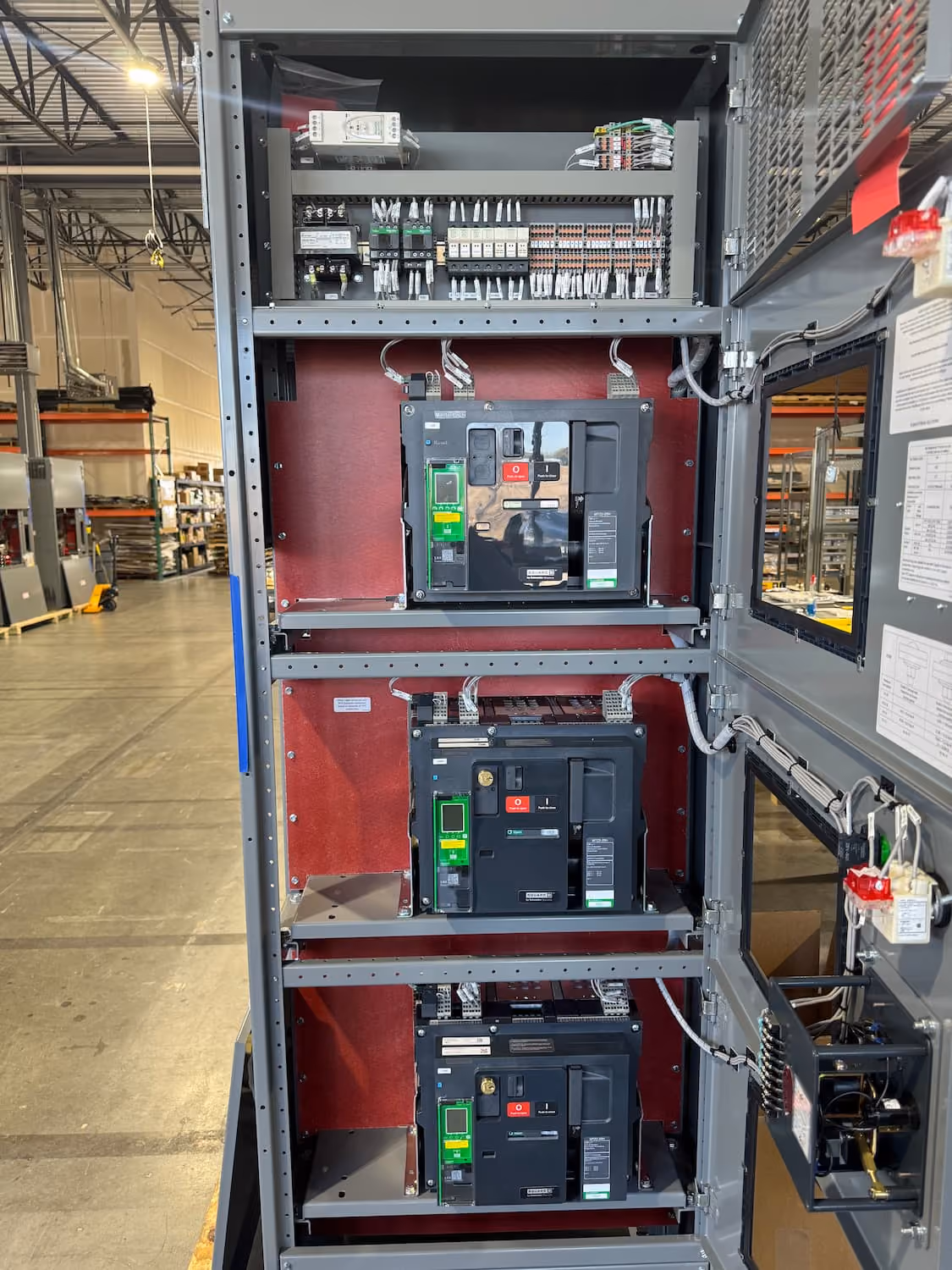 Open electrical panel inside an industrial warehouse with three large circuit breakers mounted vertically, connected with wiring and components, and a visible interior structure.