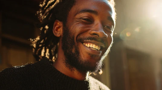 Smiling man with dreadlocks and beard, lit by warm sunlight from the right.