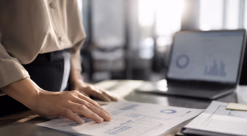 Person reviewing printed charts and graphs with a laptop displaying similar data in the background.