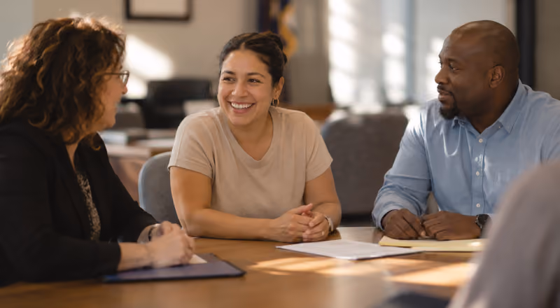 Three diverse professionals smiling and talking around a table with papers in a meeting room.
