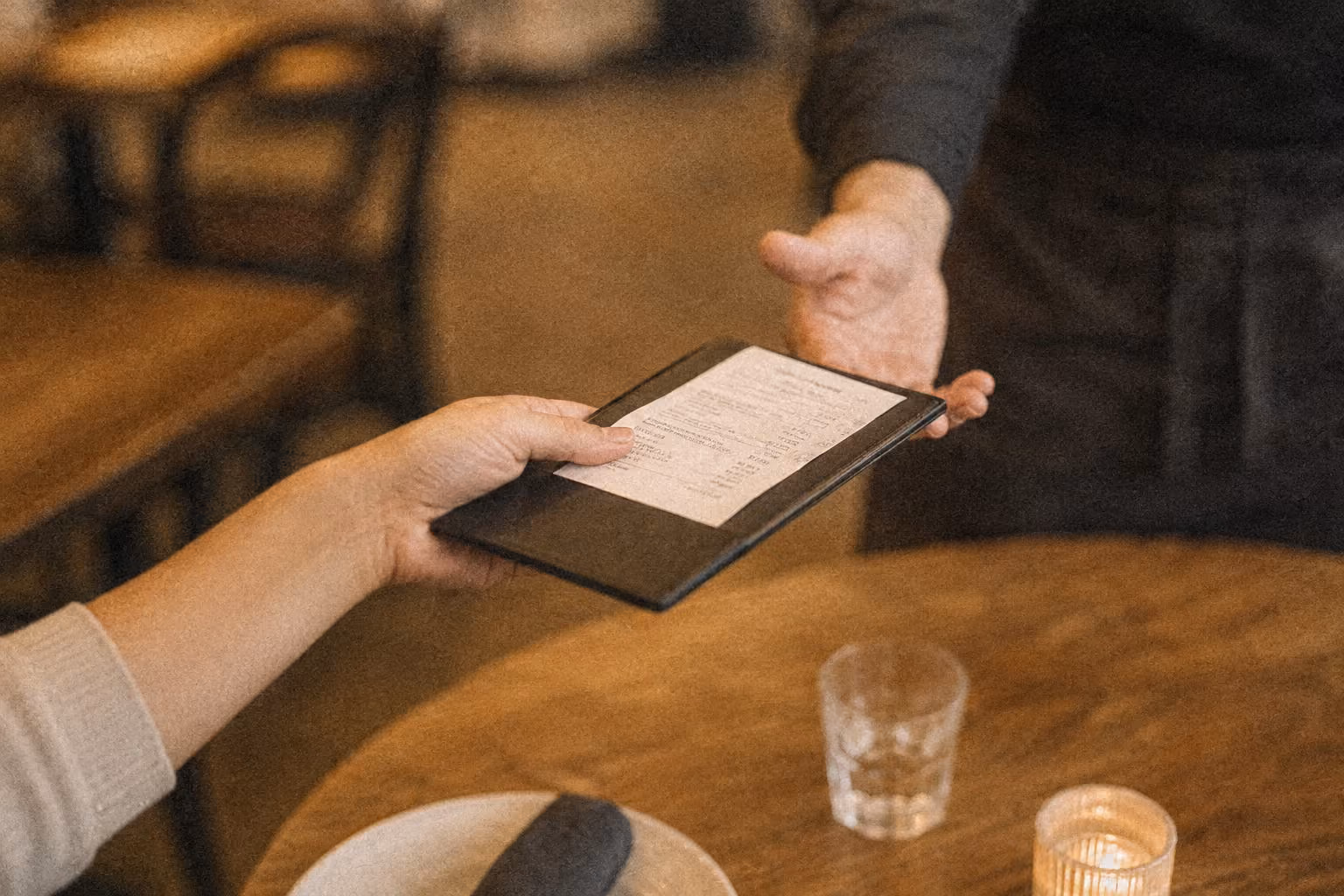 A diner hands a bill in a black folder to a waiter over a wooden table with a glass and candle.