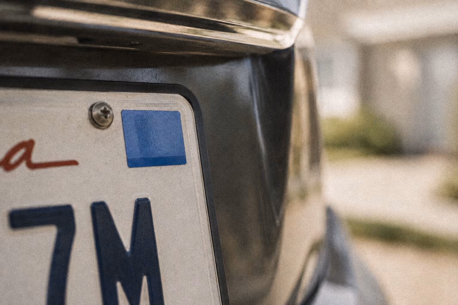 Close-up of a car license plate with a blue sticker on the top right corner and partial characters visible.