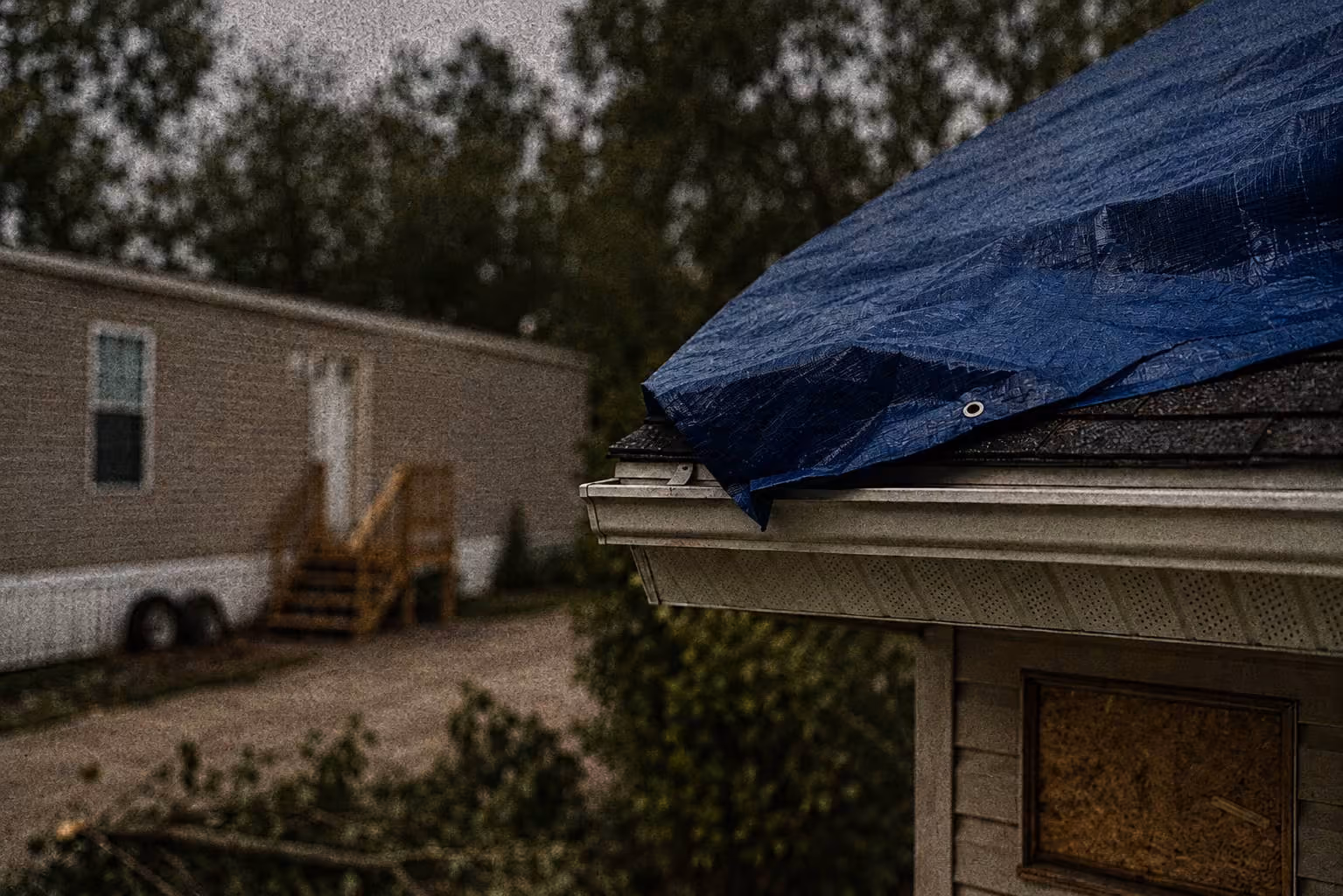 Blue tarp covering the roof corner of a building with boarded window; trailer home in blurred background.