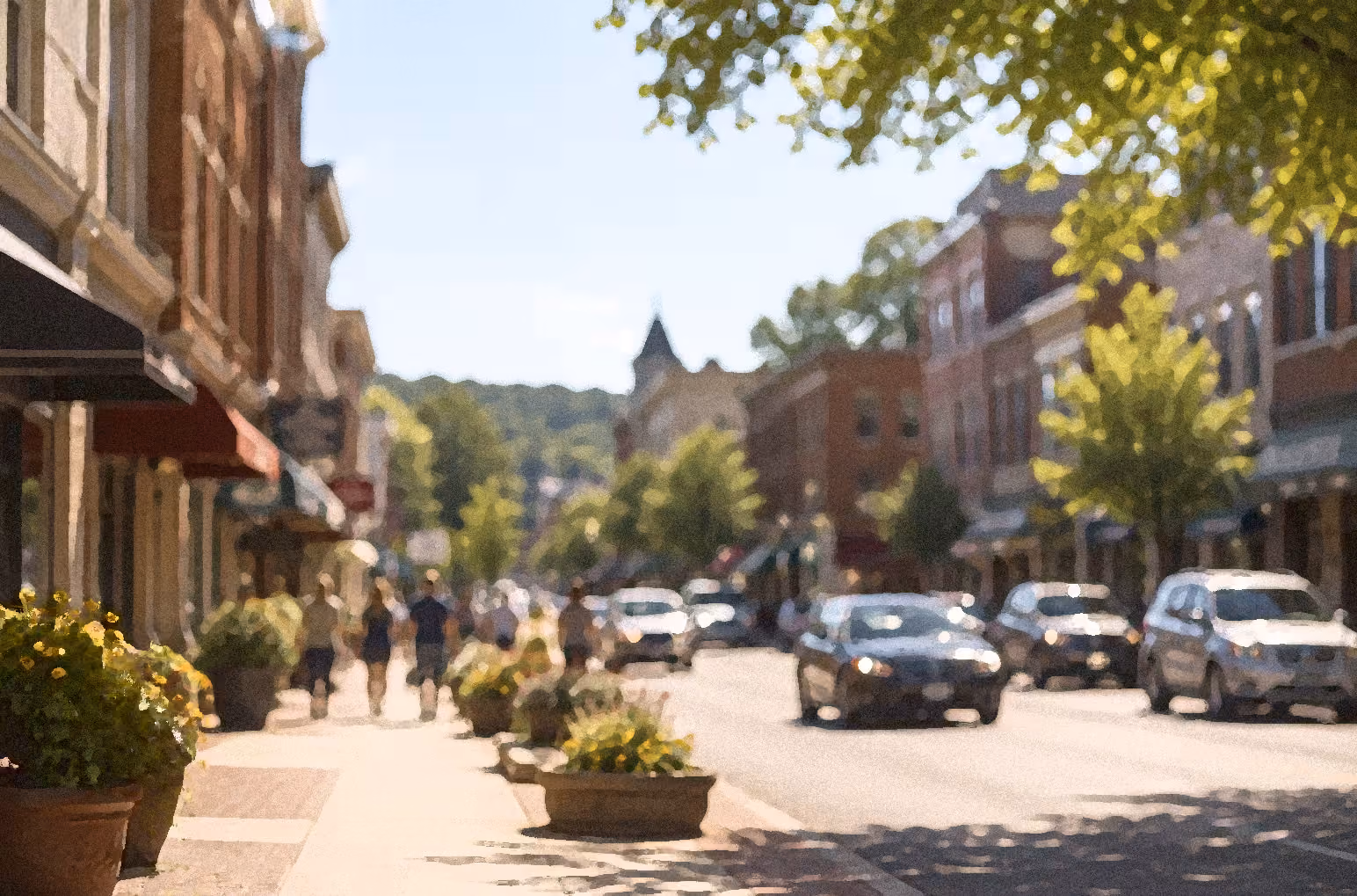 Sunny small-town street with brick buildings, parked cars, pedestrians, and potted plants along the sidewalk.