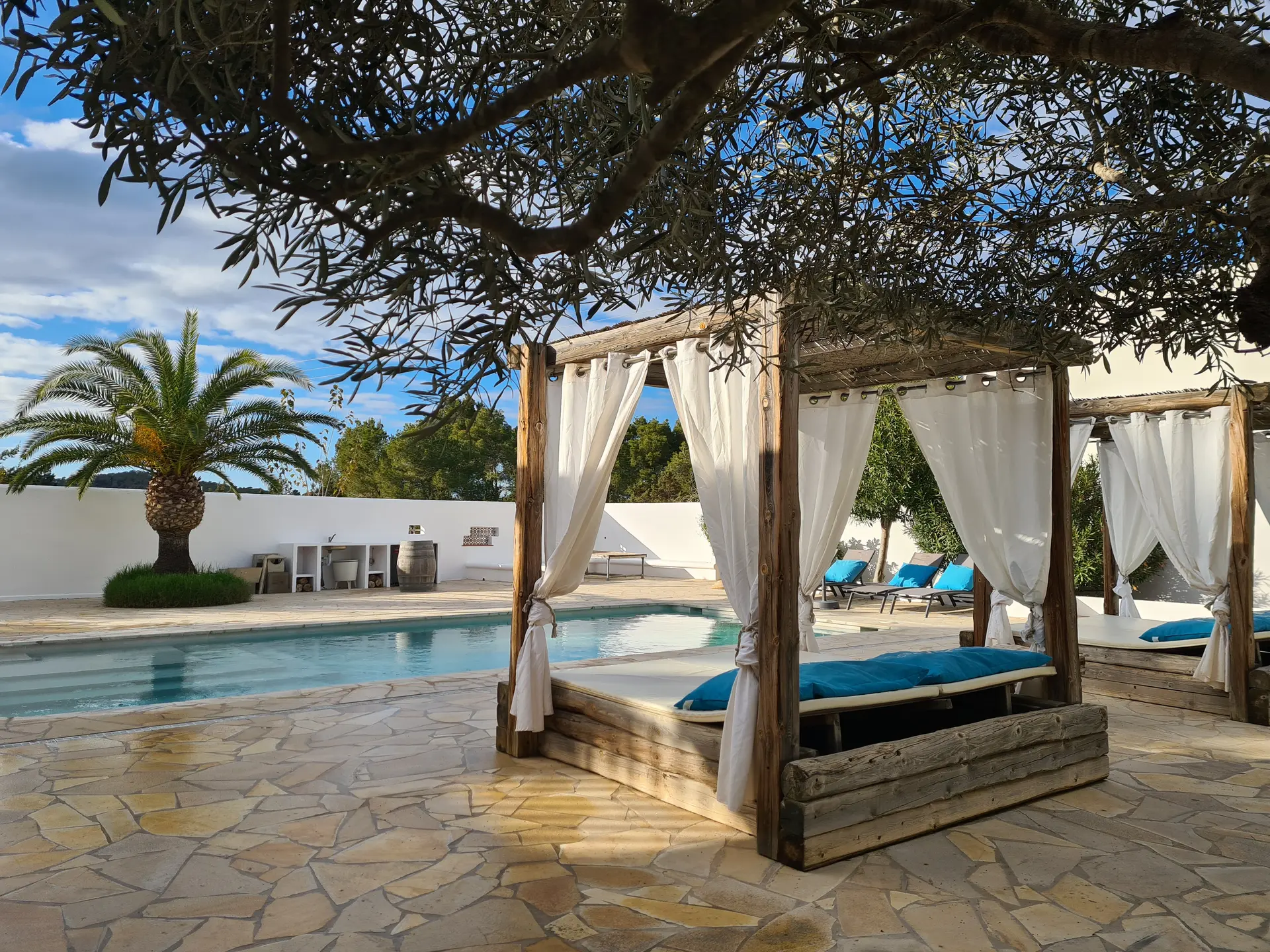 Outdoor pool area with a wooden pergola bed featuring white curtains and blue cushions, surrounded by stone tiles, palm trees, and sun loungers.