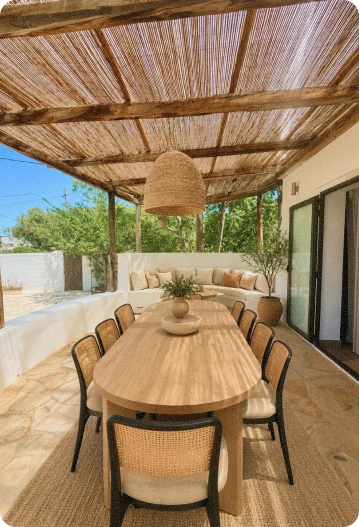 Outdoor dining area featuring a long wooden table with eight chairs under a pergola made of wooden beams and reed shades, with a hanging woven pendant light and a curved white bench with neutral cushions along the wall.