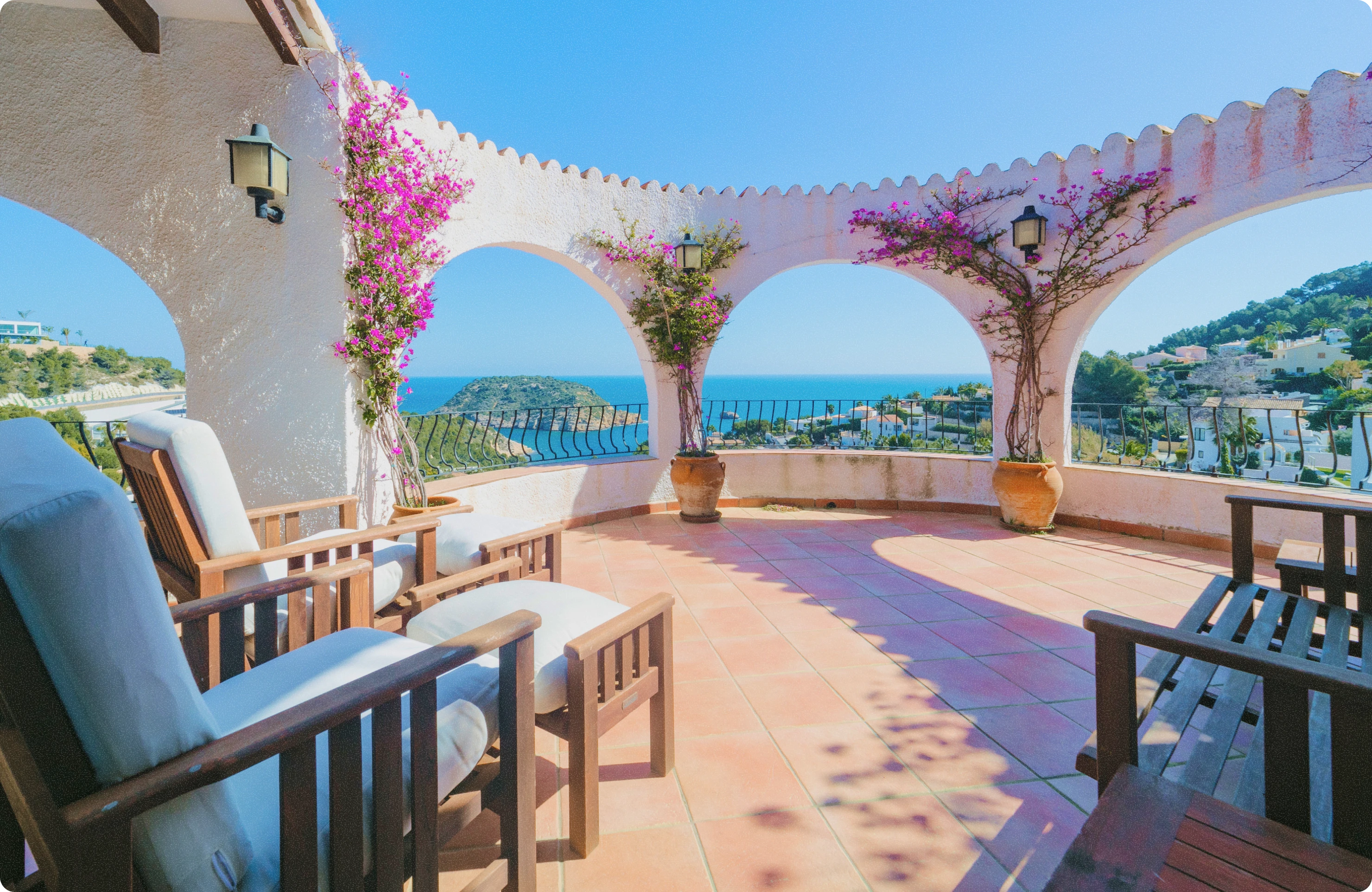 Terrace with wooden lounge chairs and a bench under white arches decorated with purple flowering vines, overlooking a coastal view with blue sea and green hills.