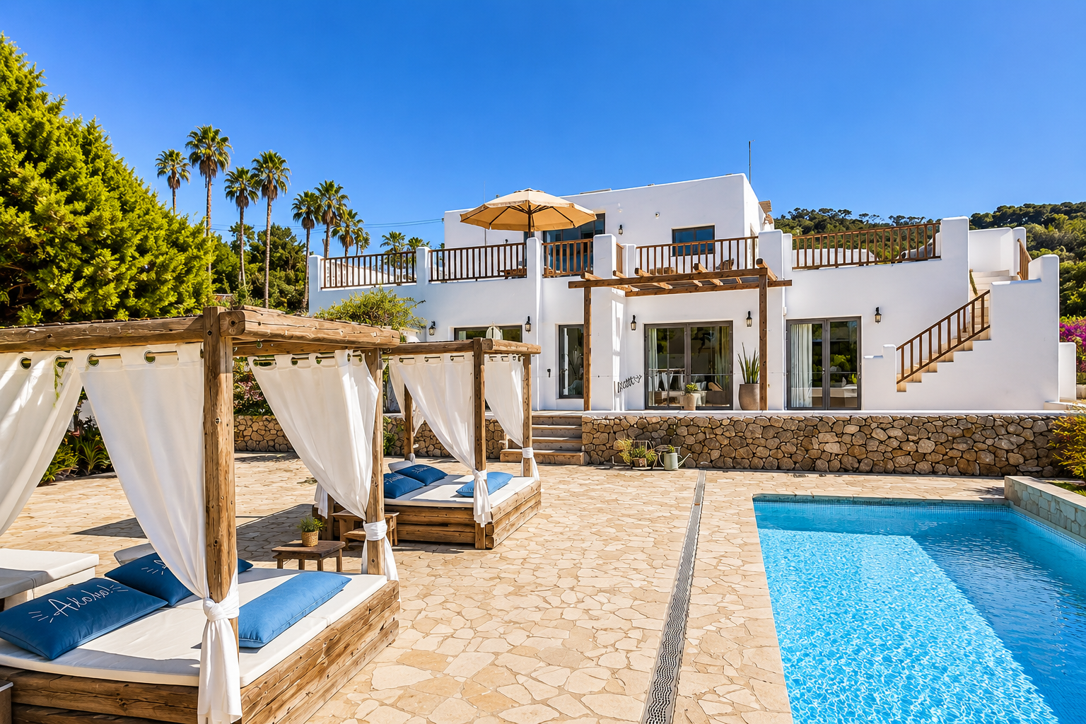 Modern white villa with upper terrace and wooden railing, stone patio with two wooden canopy daybeds with white curtains and blue cushions, small side tables, and a sparkling rectangular swimming pool under a clear blue sky.