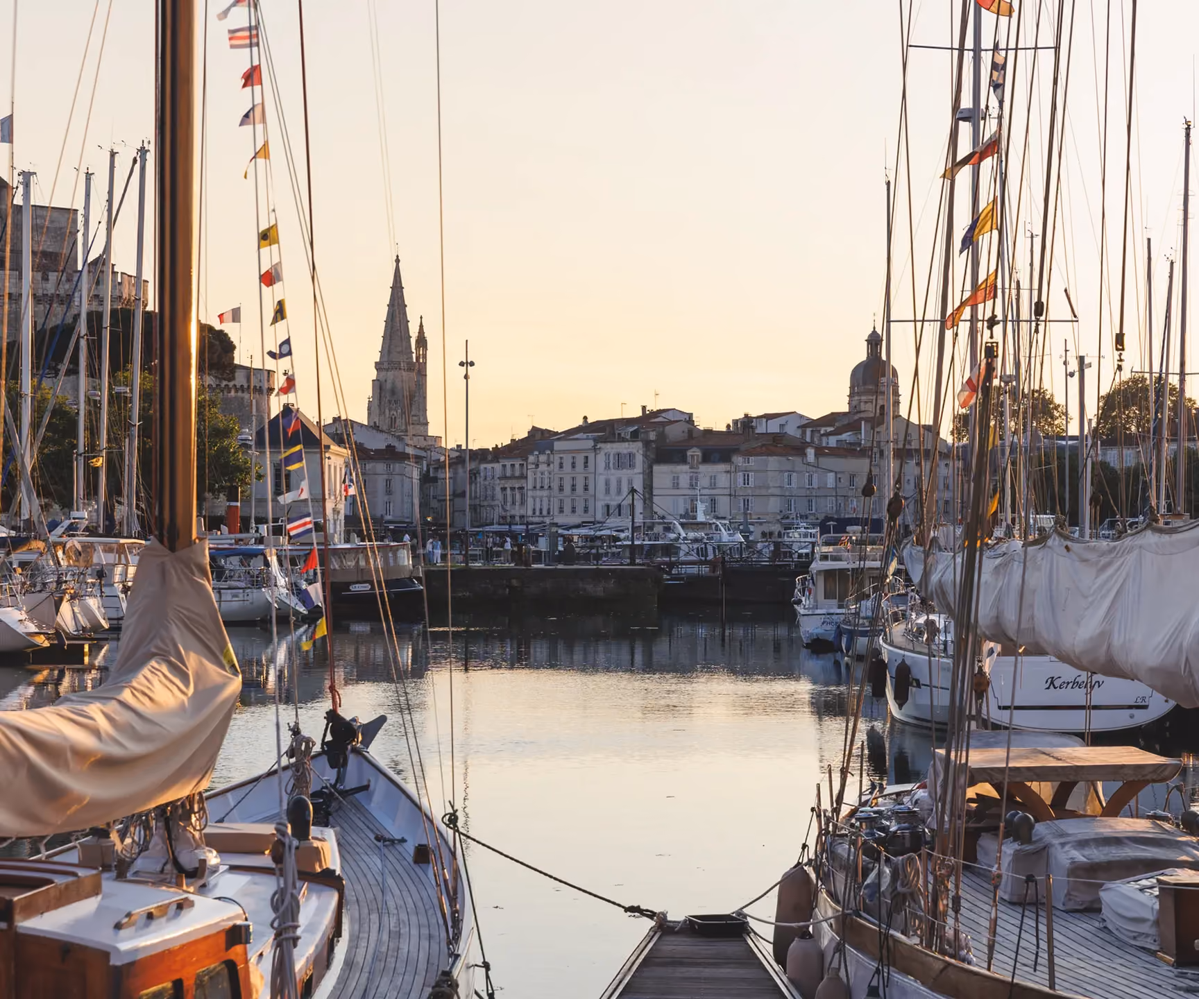 Sailboats docked at a marina with a calm water view of old buildings and church towers at sunset.