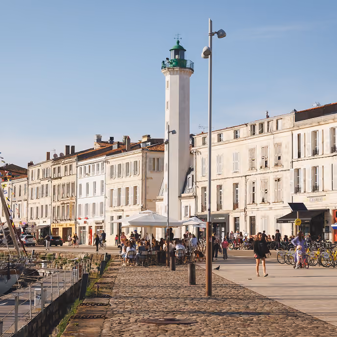 Tall white lighthouse with green top among seaside buildings and people dining outdoors on a sunny day.