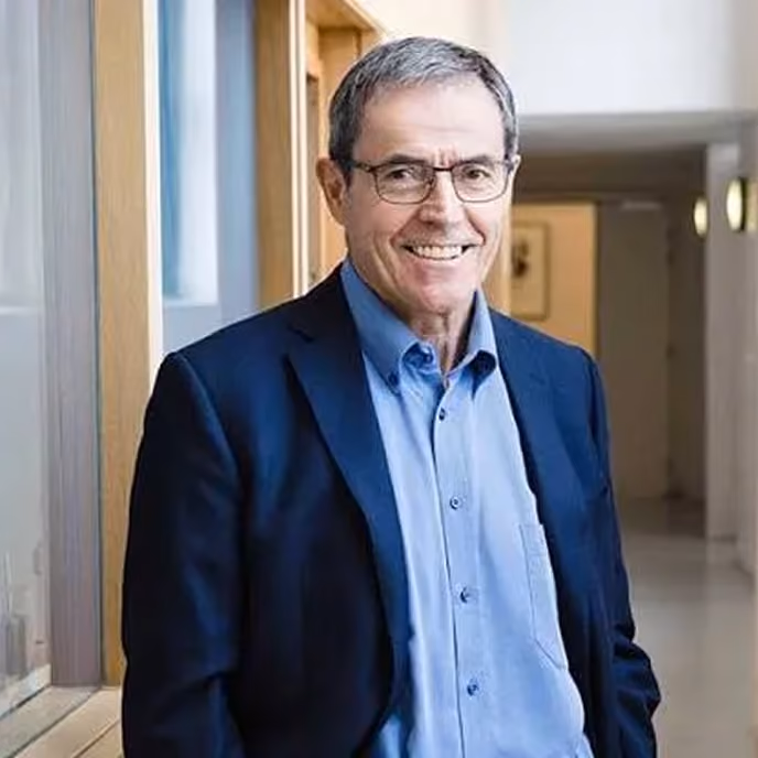 Smiling middle-aged man wearing glasses, blue shirt, and navy blazer in an indoor hallway.