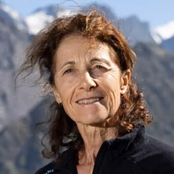 Woman with curly brown hair outdoors with mountains and blue sky in background.