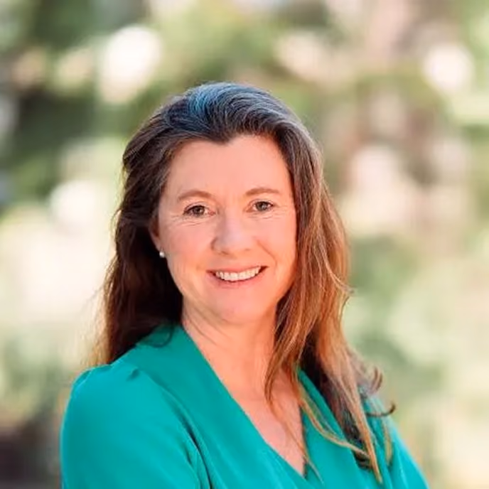 Smiling woman with long brown hair wearing a green top with a blurred natural background.