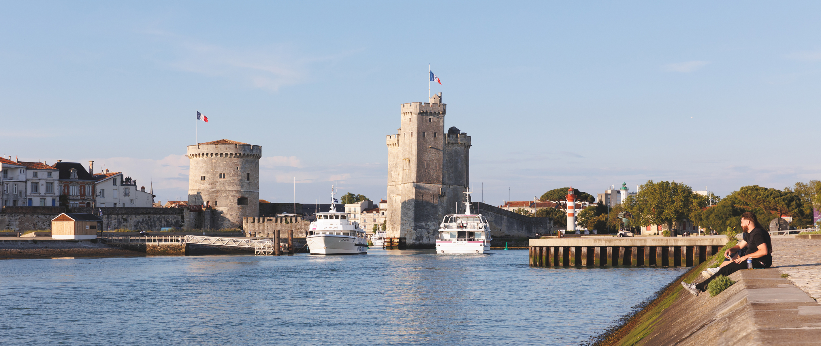 Two boats passing between two stone towers with French flags at a waterfront harbor in sunlight.