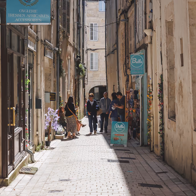 Sunlit narrow street with people walking and standing near shops and flower decorations.