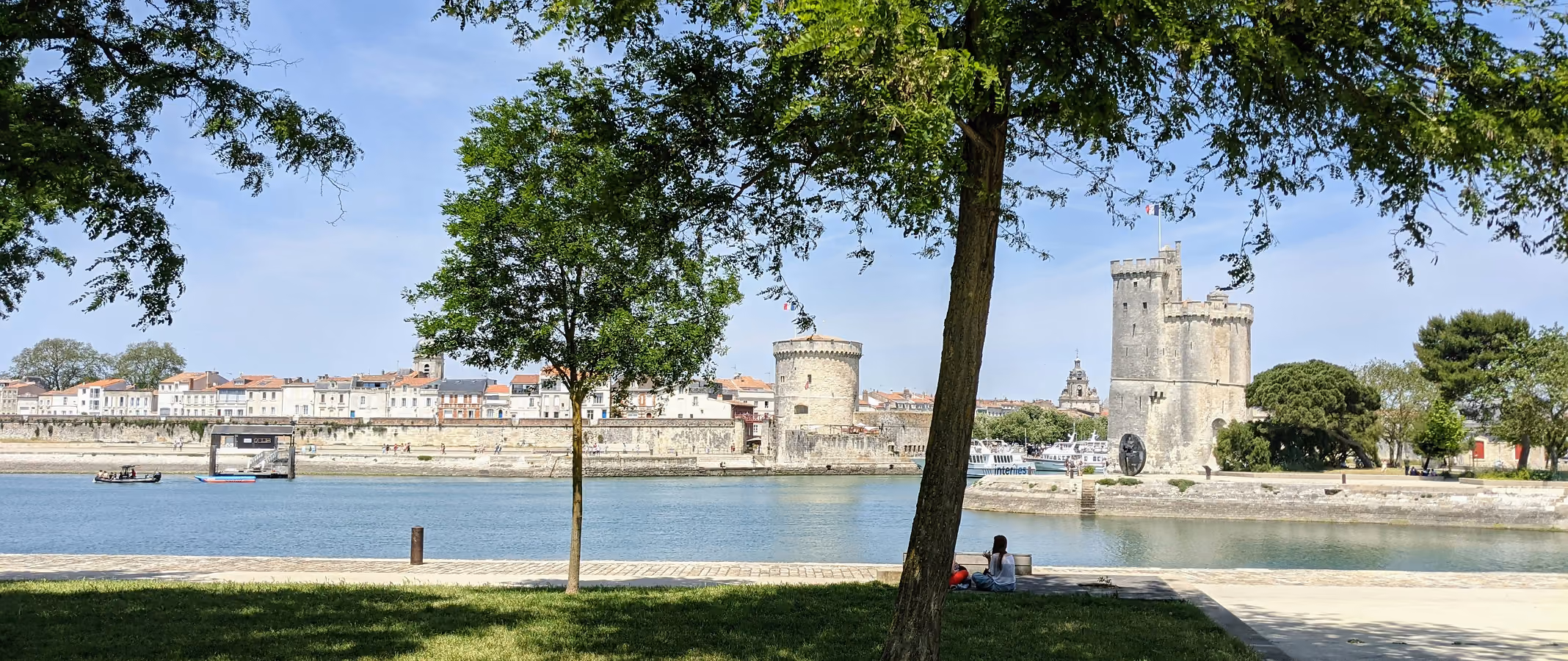 Riverside view with trees, people sitting on grass, stone towers, and buildings across the water on a sunny day.