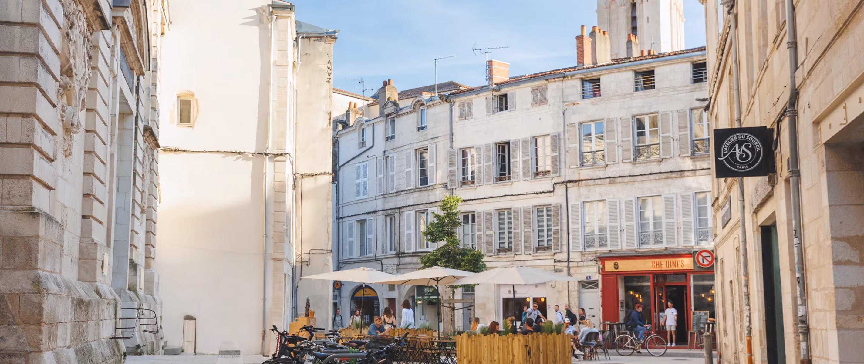 Outdoor cafe scene with people sitting under umbrellas by bicycles in front of historic white stone buildings on a sunny day.
