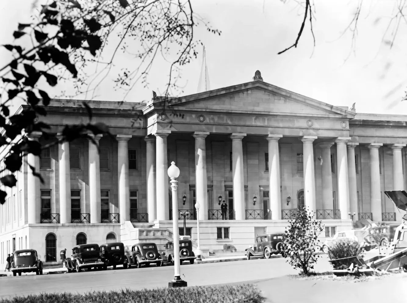 1930s black-and-white view of a classical columned civic building with vintage cars parked out front