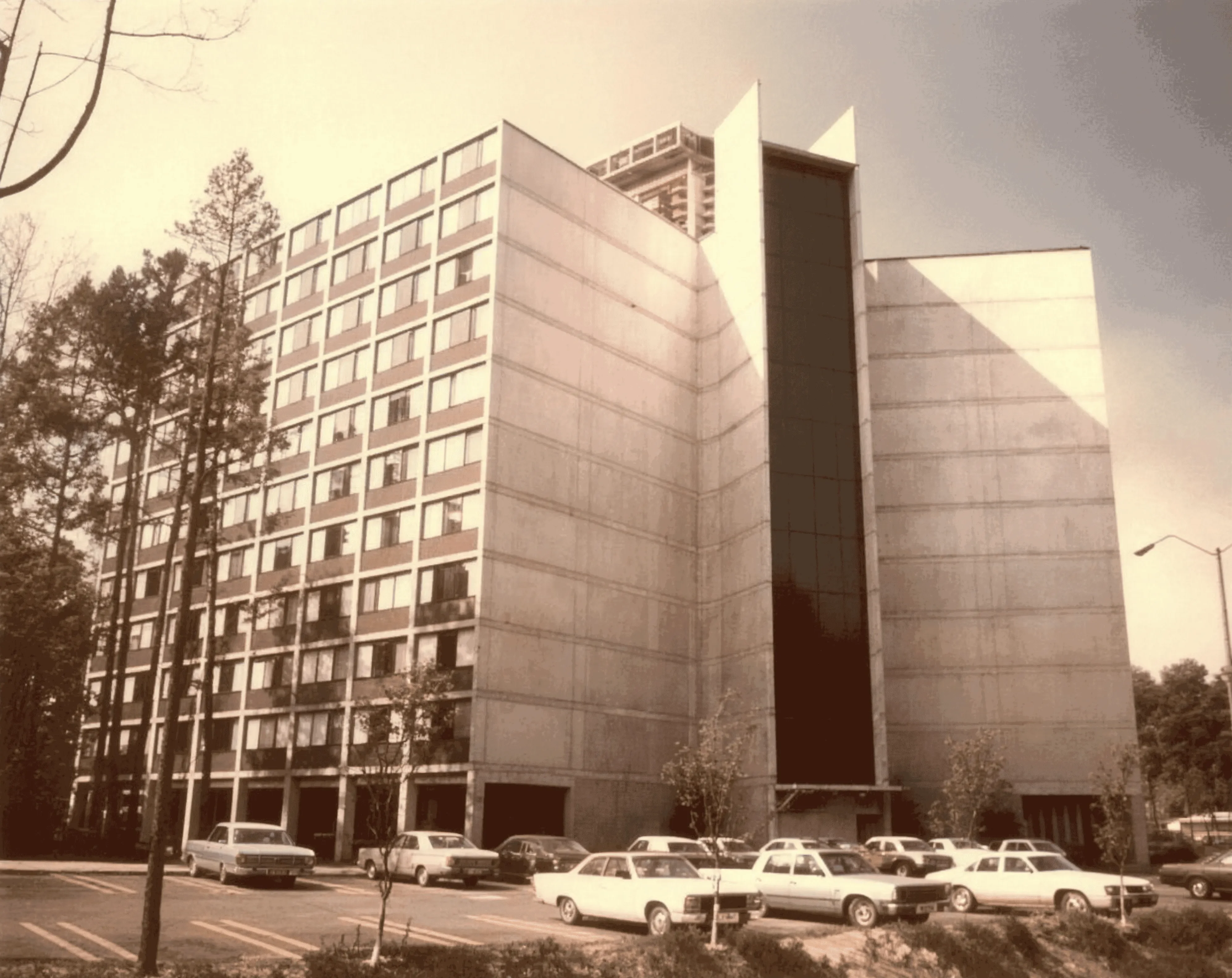 Sepia 1970s photo of a multistory concrete residential tower with cars parked in front