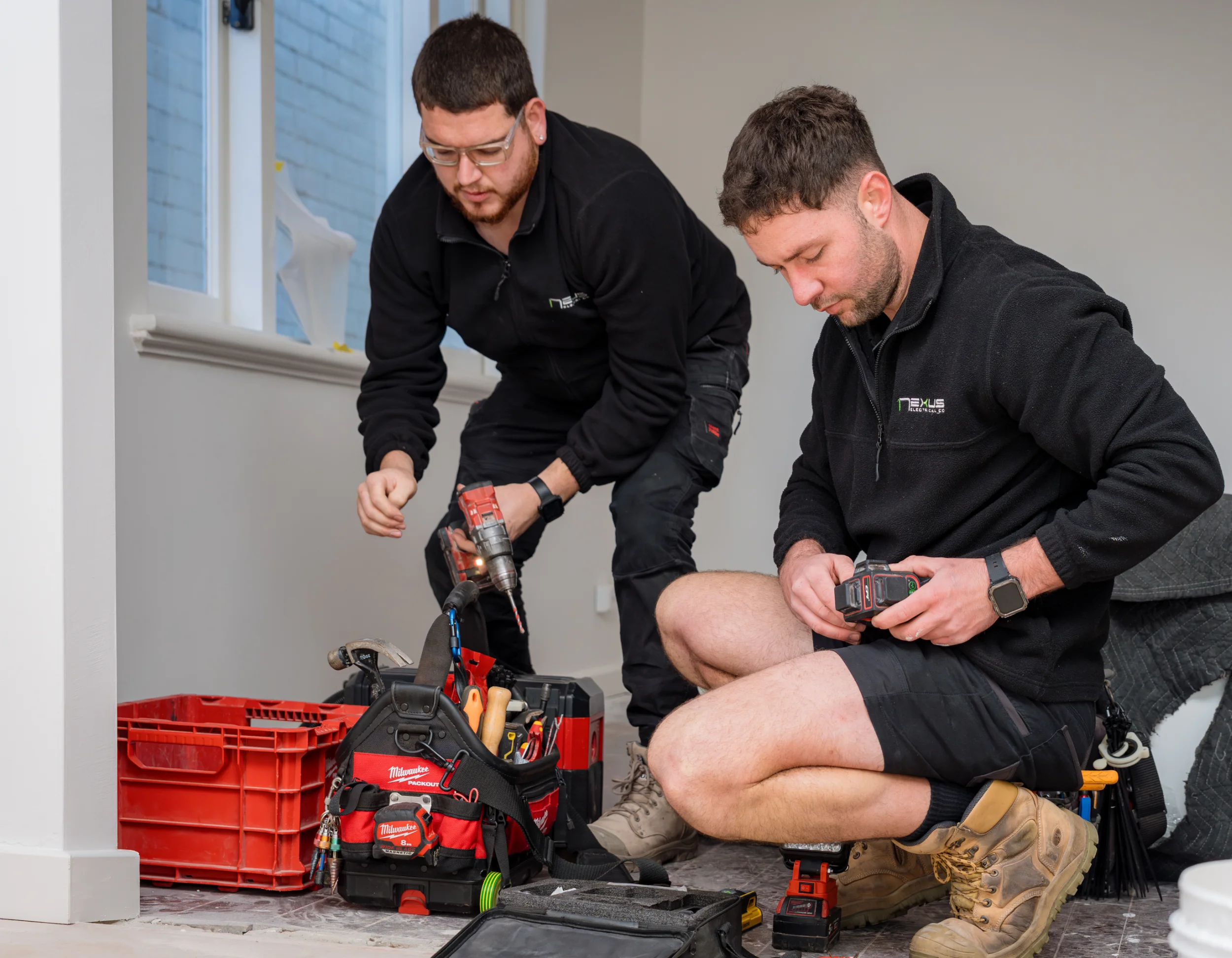 Two electricians in black uniforms kneeling indoors working with tools and power drills near a window.