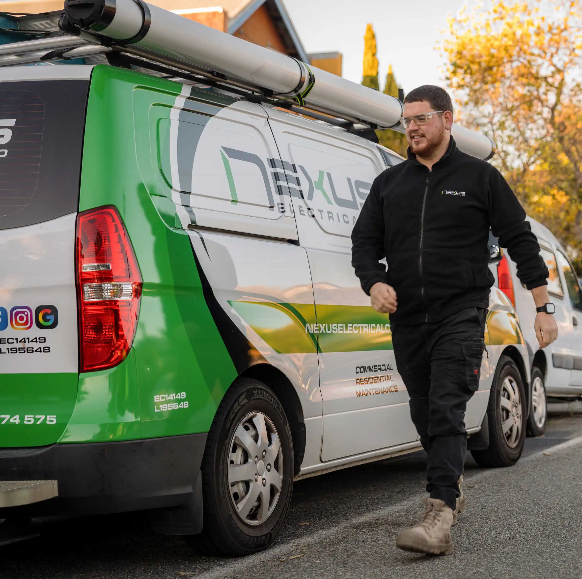 Man in safety glasses and black jacket walking beside a green and white Nexus Electrical van.
