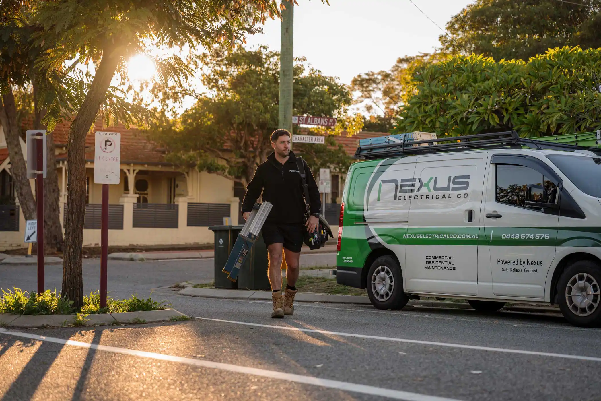 Electrician carrying tools and ladder walking near a Nexus Electrical Co van on a sunny street.