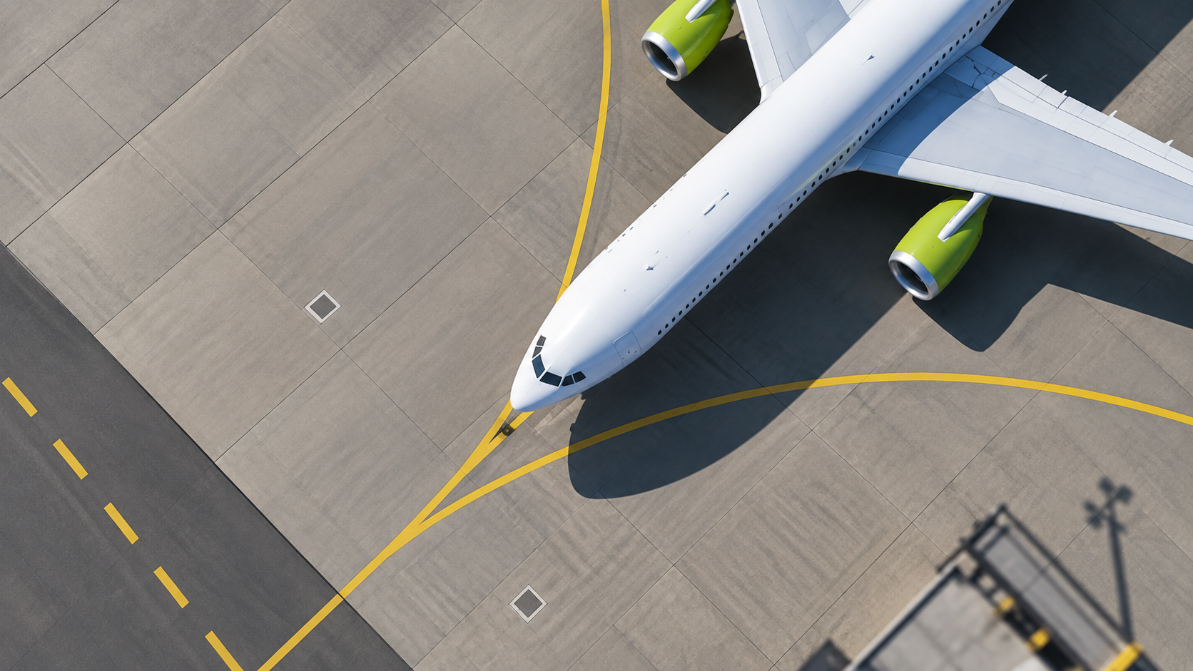 Overhead view of a commercial aircraft on an airport apron with yellow taxiway lines and green engine accents