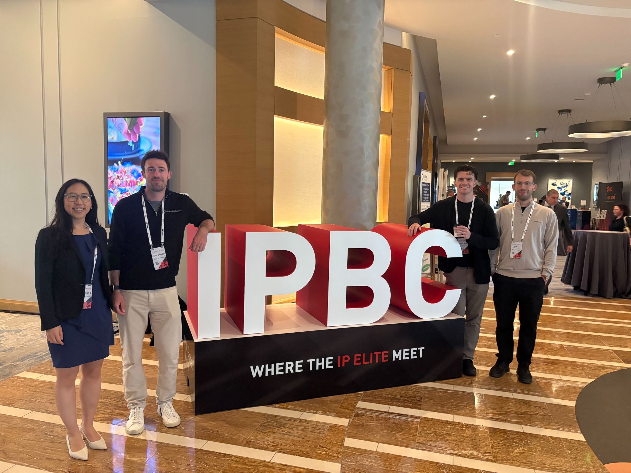 Four people posing beside a large “IPBC” sign reading “Where the IP Elite Meet” at a conference lobby