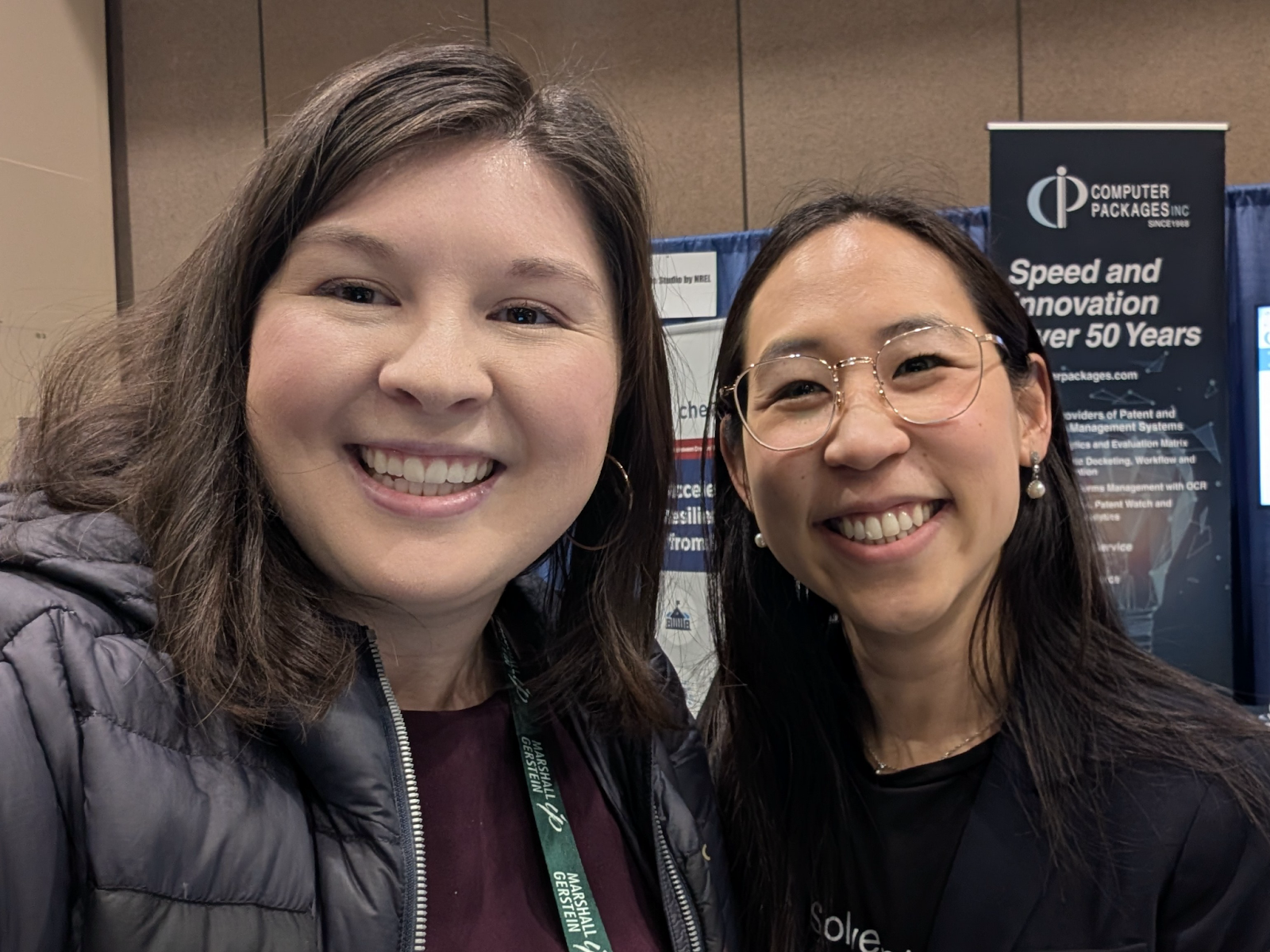 Two people smiling for a photo at the AUTM conference exhibit hall, with an exhibitor banner reading “Speed and Innovation Over 50 Years” in the background