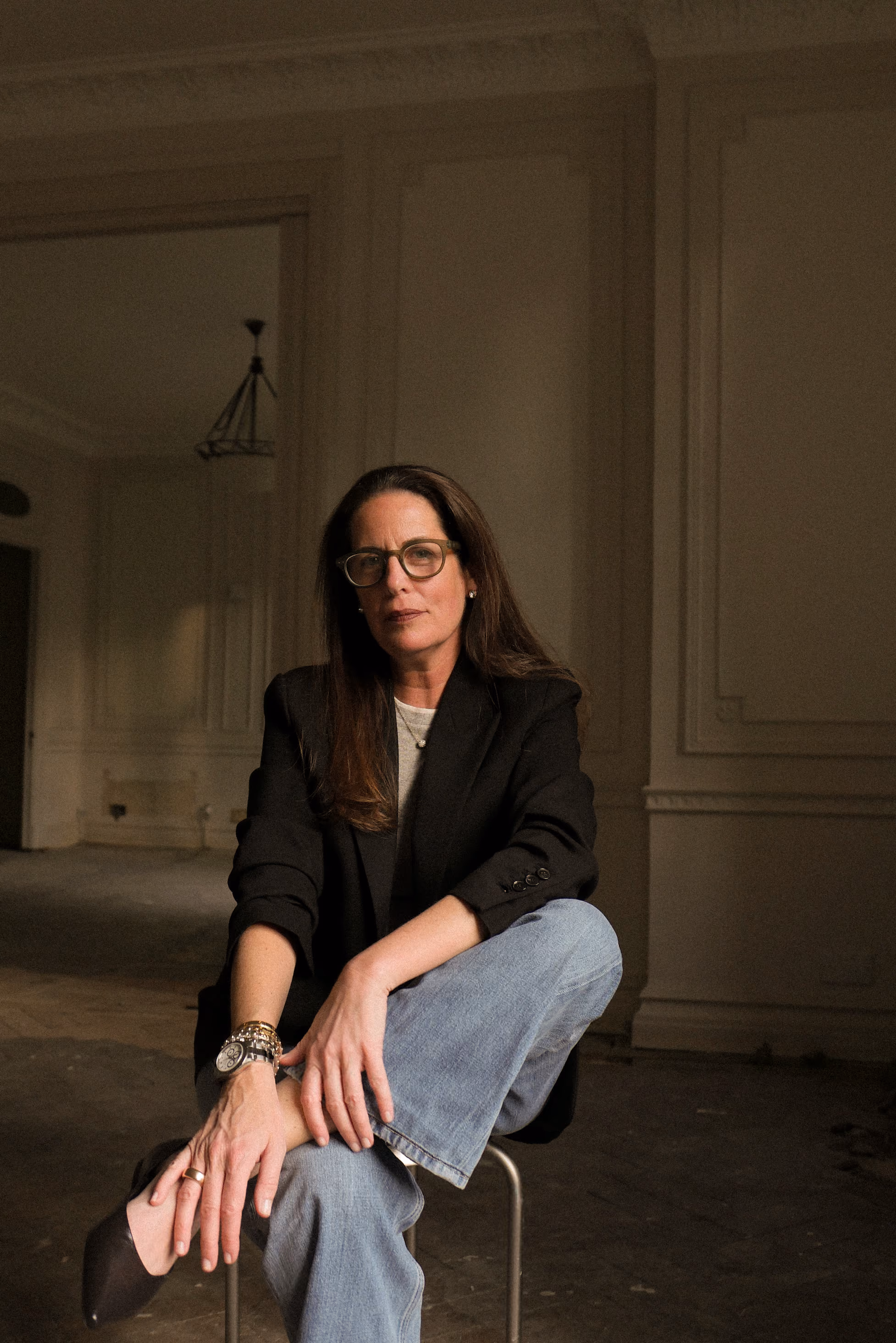 Stephanie wearing a black blazer and jeans sits on a chair in a dimly lit room.