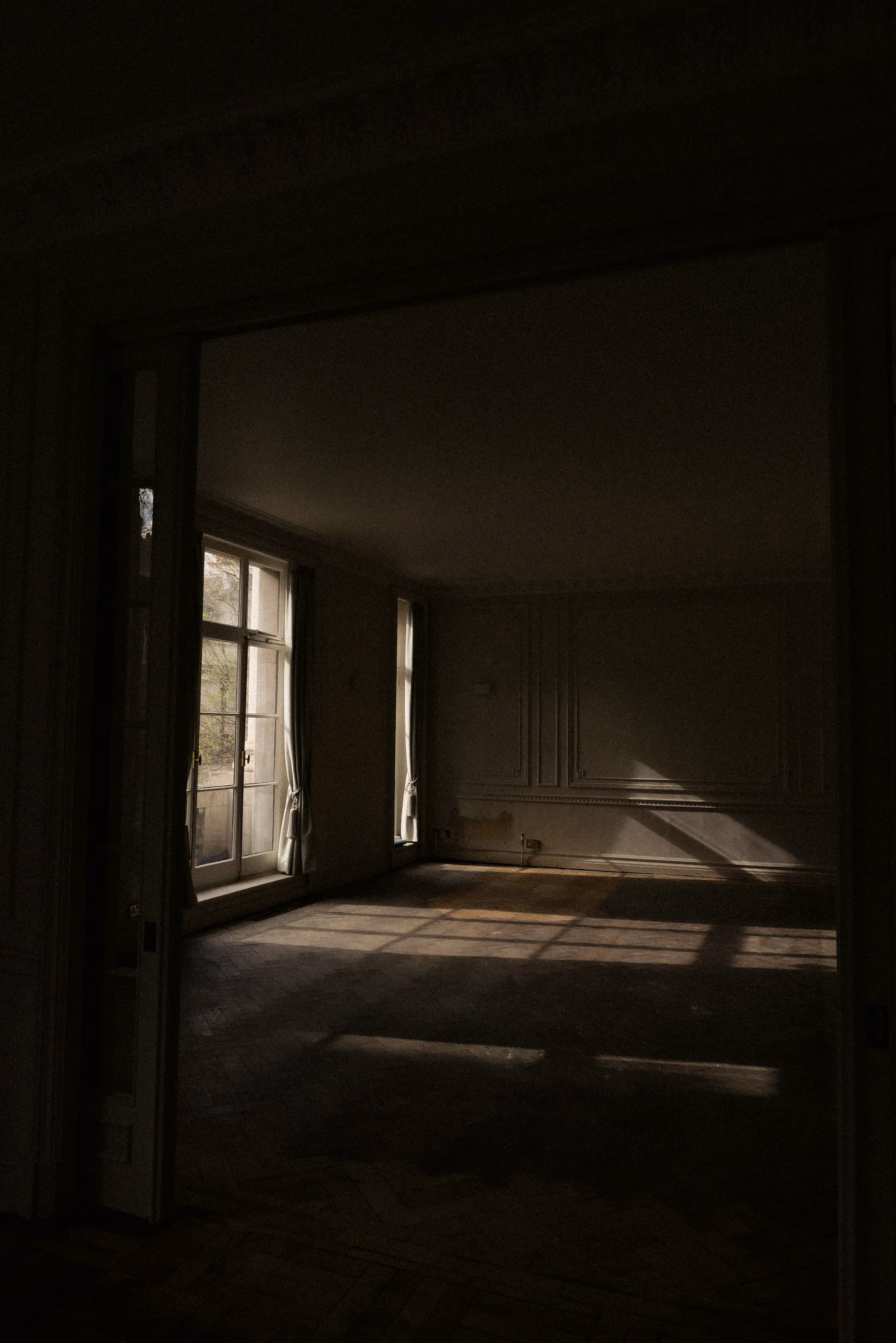Dimly lit empty room with tall windows casting light and shadows on the wooden floor.