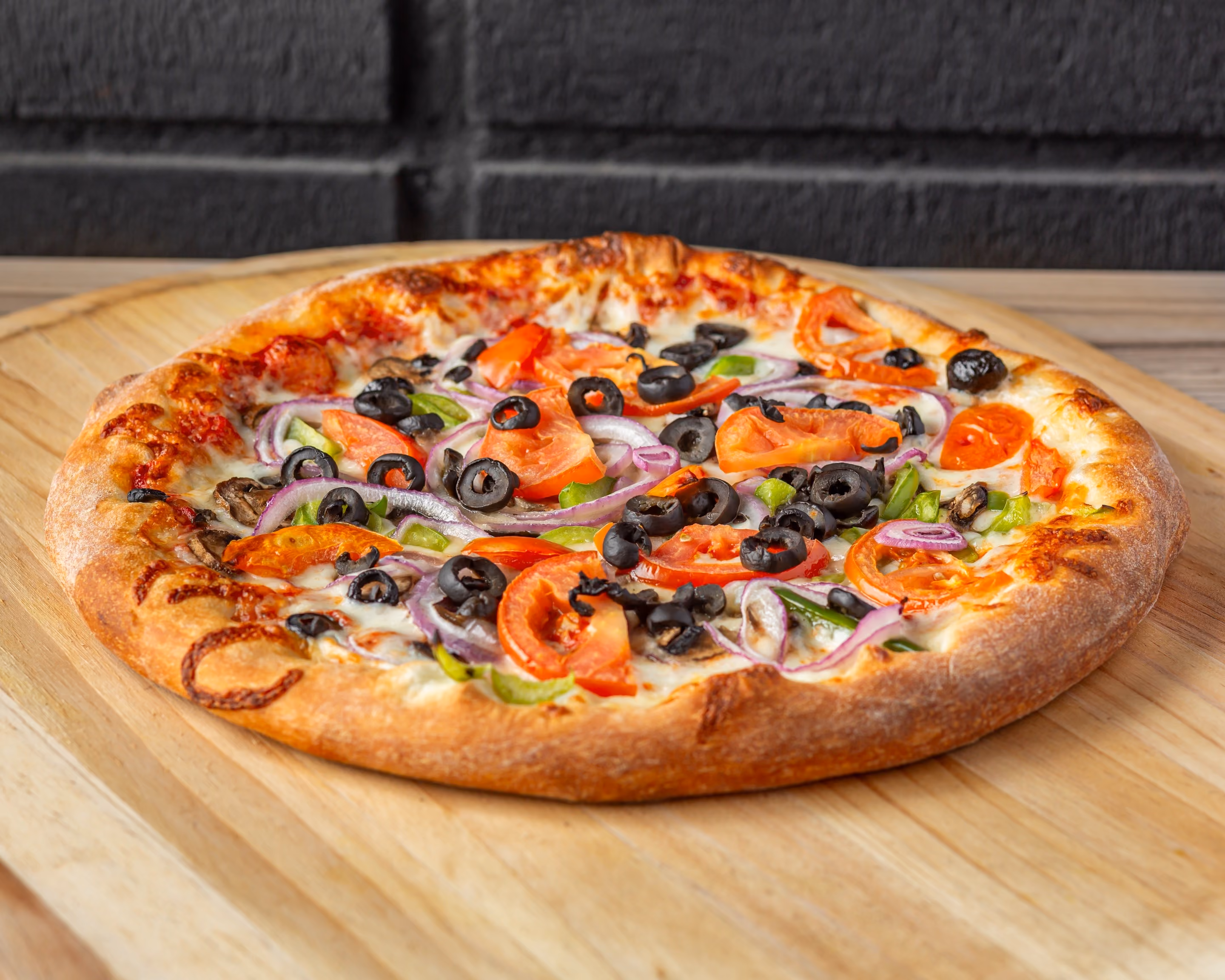 Vegetable pizza with black olives, tomatoes, red onions, and green peppers on a wooden board.