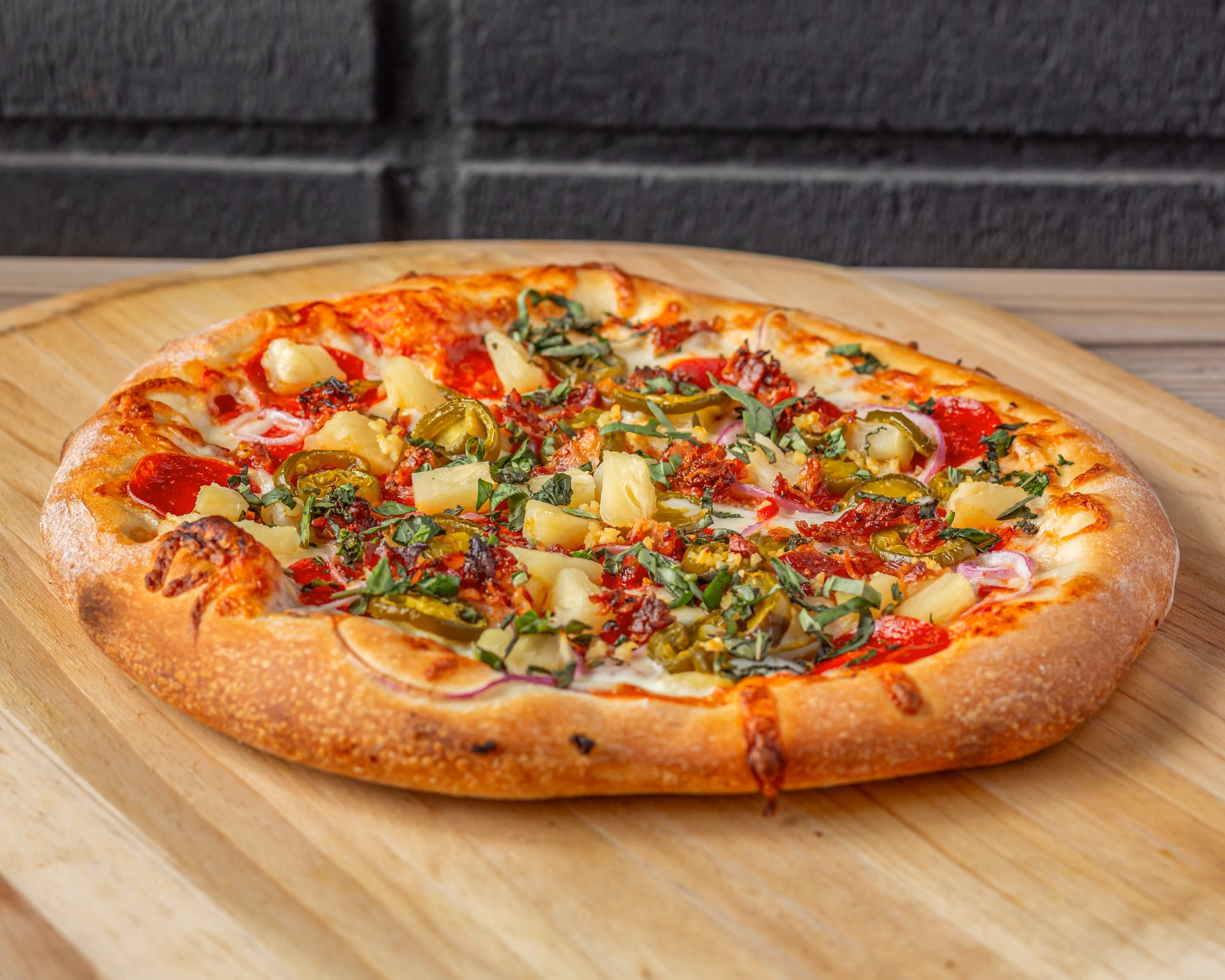 Pizza with pepperoni, pineapple, jalapeños, basil, and cheese on a wooden board.