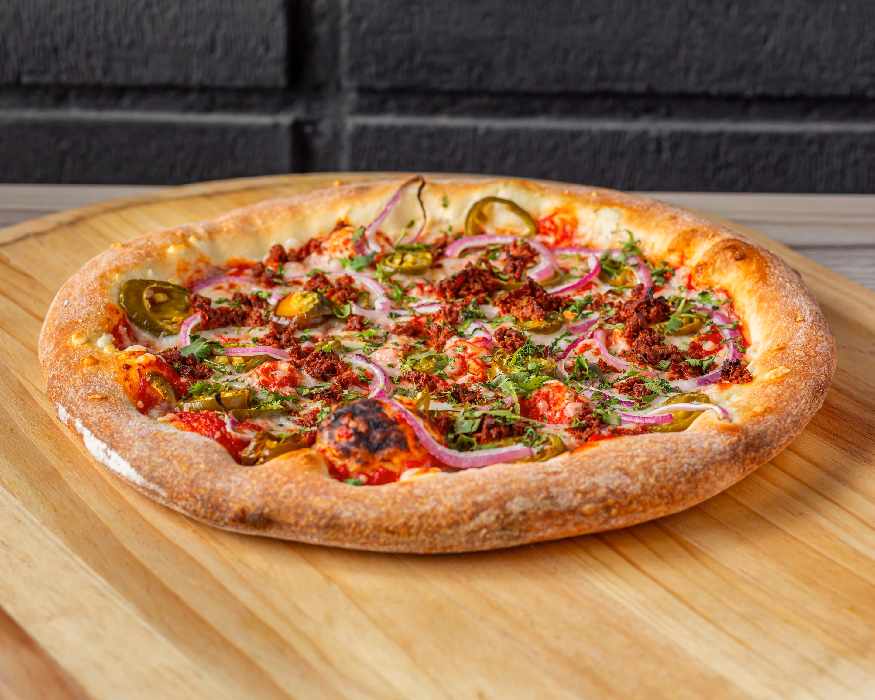Fresh pizza topped with jalapeños, red onions, ground meat, and chopped herbs on wooden board.