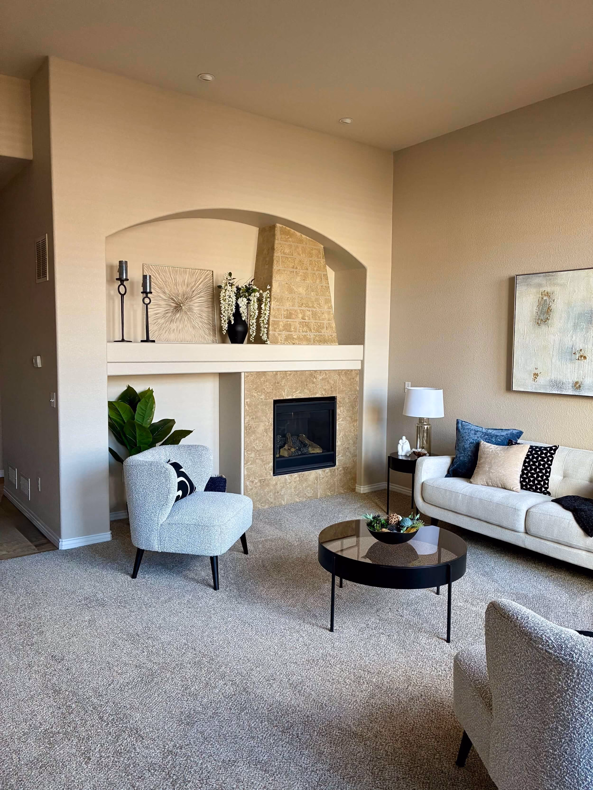 Modern living room with light beige walls, a stone fireplace with a recessed alcove above decorated with tall candles and flowers, a white sofa with blue and patterned cushions, two light gray armchairs, round black coffee table, side table with lamp, and a large green plant.