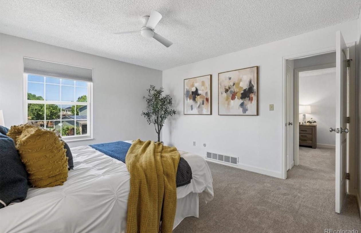 Bright bedroom with white walls and beige carpet featuring a bed with white bedding, blue and mustard yellow pillows, and a mustard yellow blanket draped on the side. There is a large window with a view of trees and houses, two abstract paintings on the wall, a green potted plant in the corner, a white ceiling fan, and an open door leading to a room with a wooden dresser and a lamp.