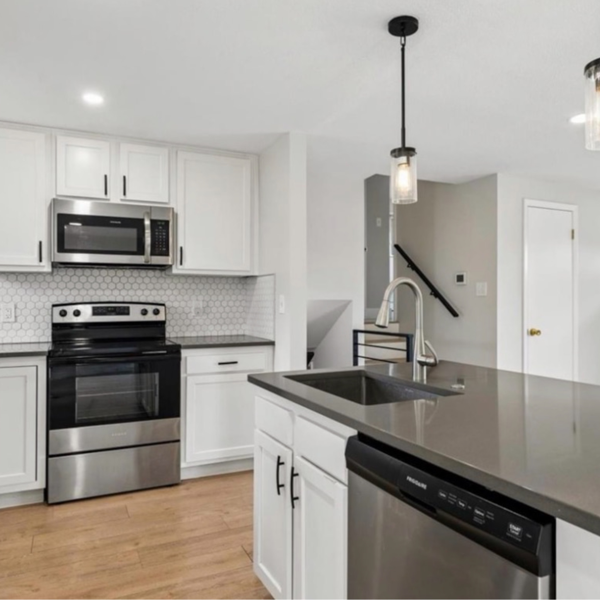 Modern kitchen with white cabinets, stainless steel microwave, oven, and dishwasher, gray countertops, hexagonal tile backsplash, wooden floor, and pendant lights.