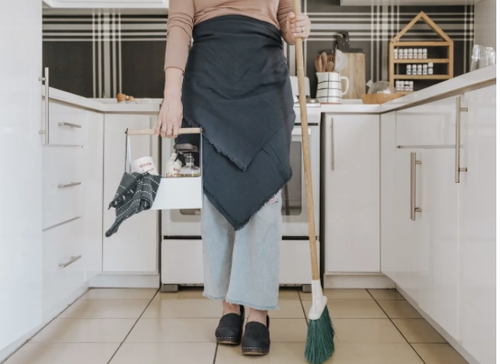 Person wearing light blue jeans, black shoes, and a dark apron standing in a kitchen holding a green broom in one hand and a small wooden basket with cleaning items in the other.