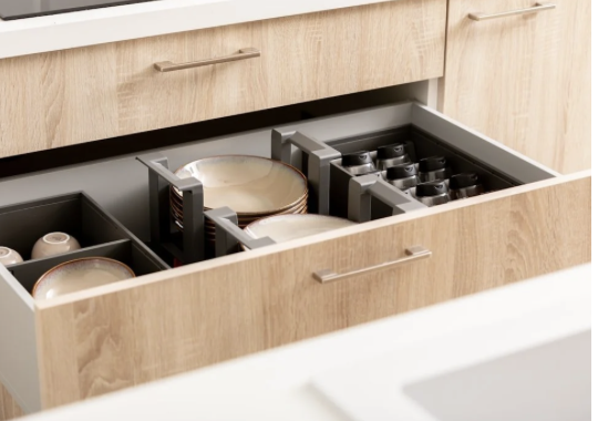 Open wooden kitchen drawer with organized compartments holding stacked plates and several black small cups neatly arranged.