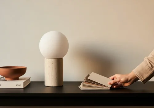 A modern minimalist black shelf with a wooden bowl on two stacked books on the left, a wooden lamp with a round white globe in the center, and a hand selecting color swatches from a fan deck on the right against a beige wall.