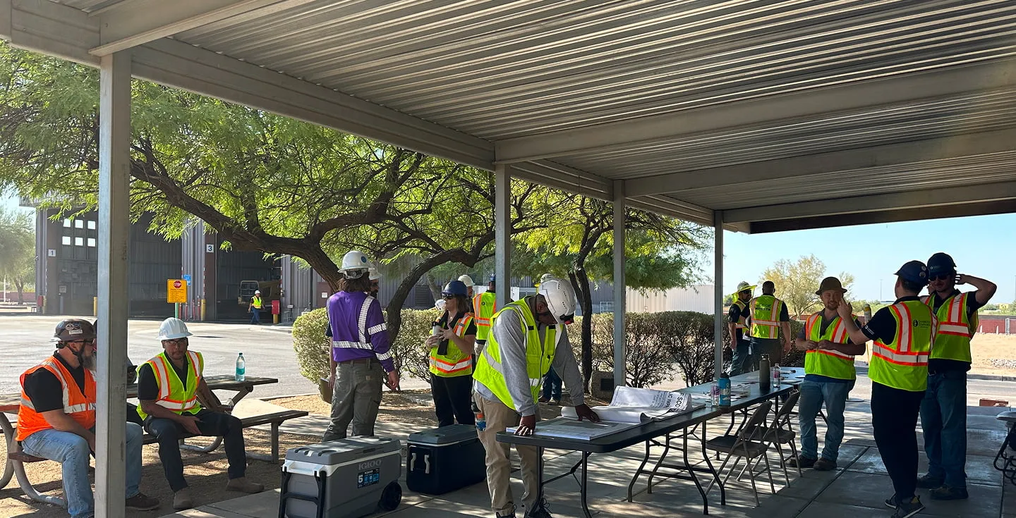 Workers seated and standing under a metal canopy during an outdoor site meeting