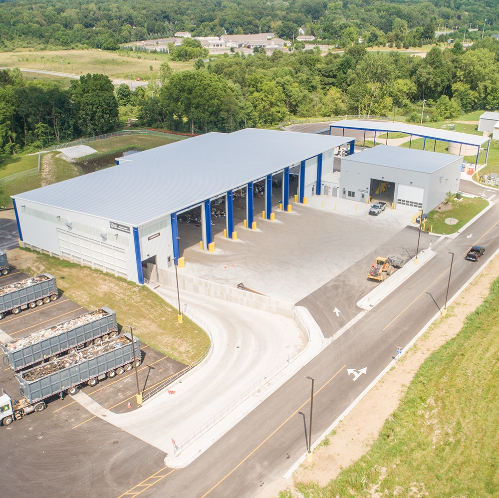 Aerial view of a county transfer station with a large building featuring multiple open bays, several trucks loaded with scrap metal parked nearby, surrounding green trees, and a paved road leading to and from the site.