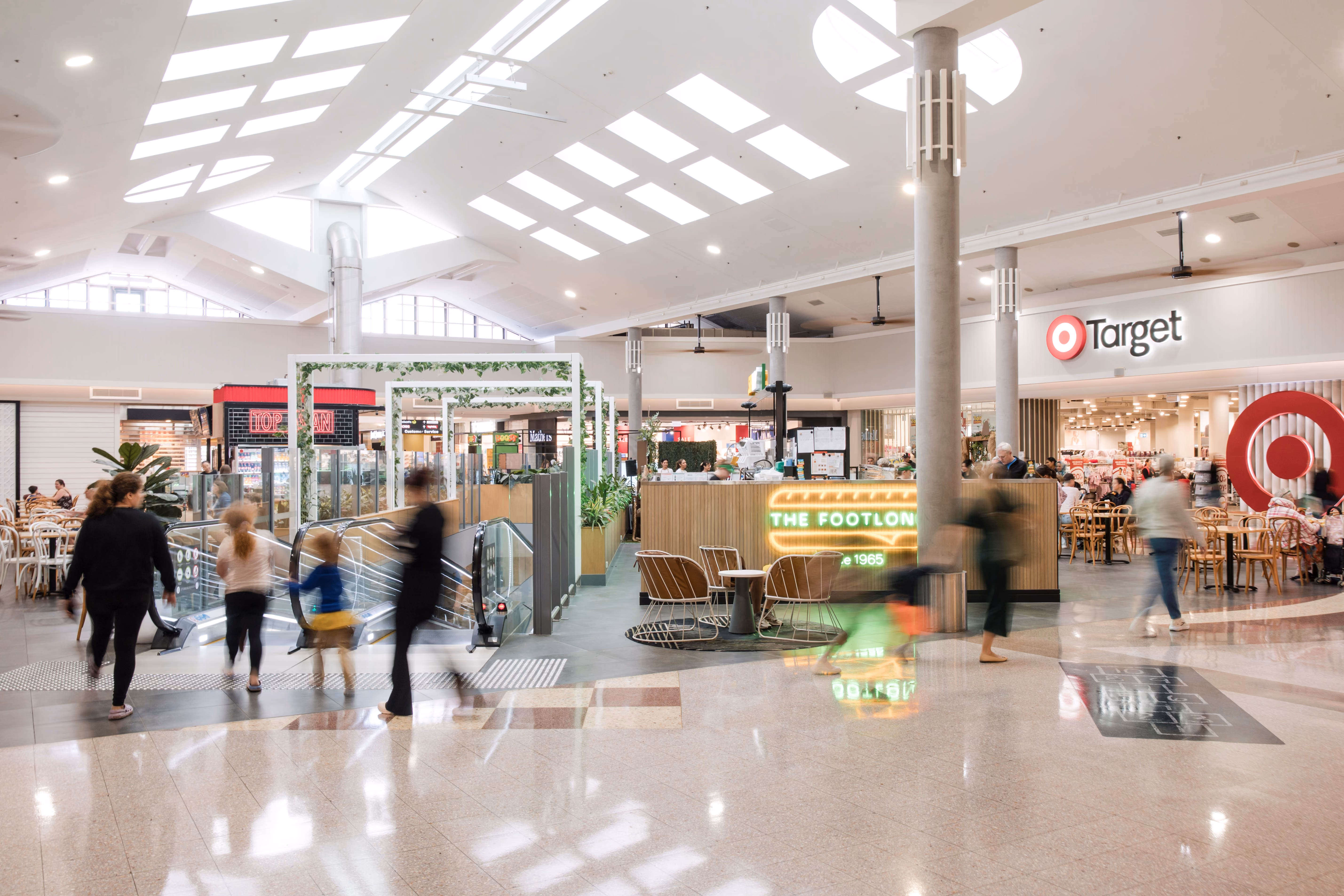 A bright, spacious shopping mall atrium with shoppers walking past escalators, seating areas, and storefronts including Target.