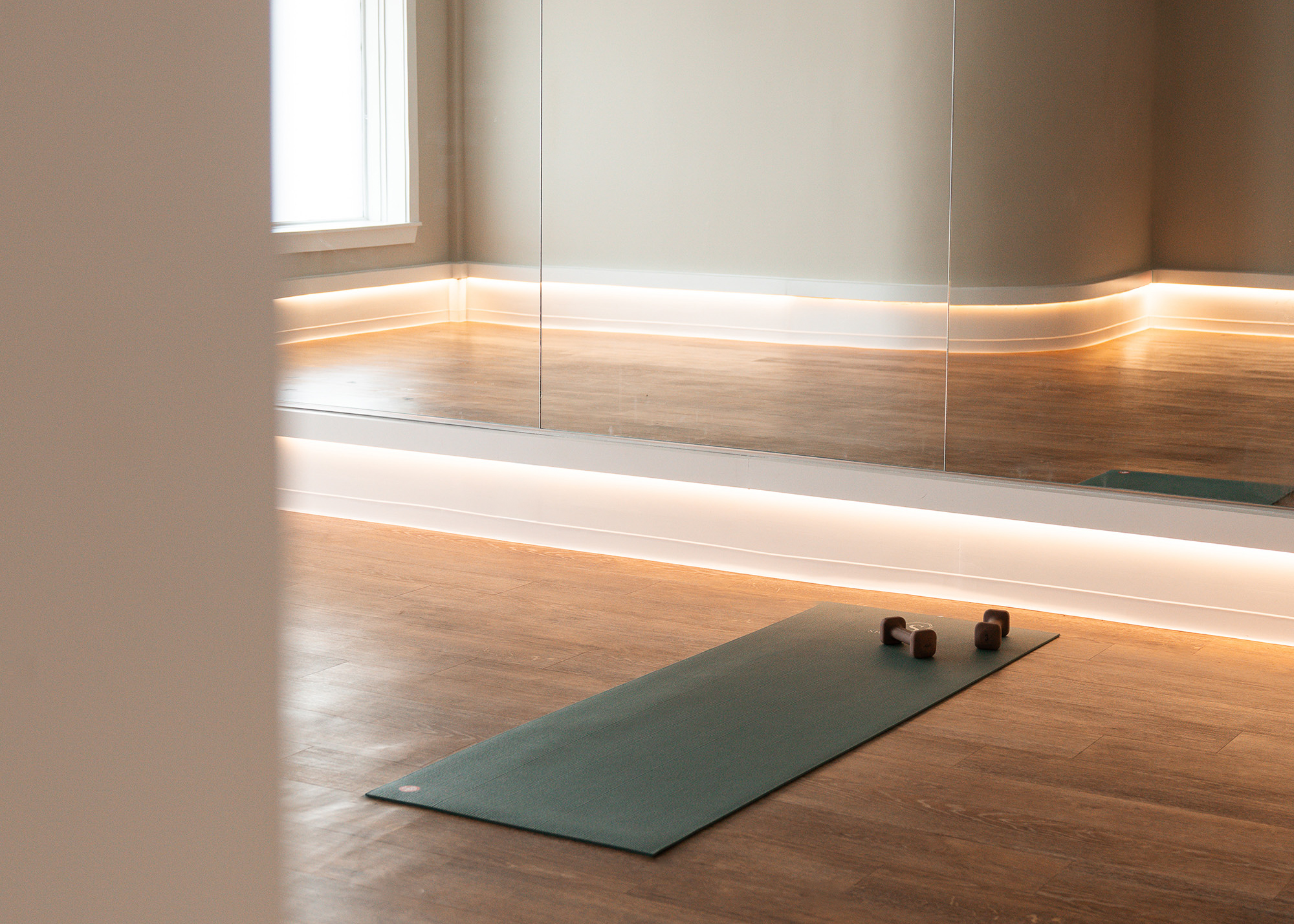 Empty fitness room with wooden floor, wall mirror, lighted baseboard, yoga mat, and two dumbbells.